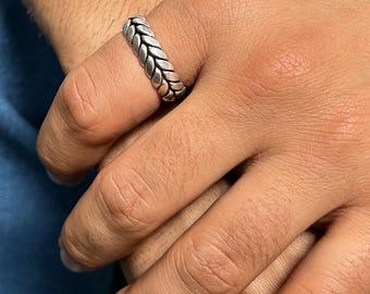 Może przedstawiać: Srebrzysty pierścionek z ciągłym, powtarzającym się wzorem warkocza lub kłosa pszenicy wokół całej obrączki. Metal ma polerowane wykończenie z przyciemnionymi wgłębieniami, które podkreślają skomplikowany wzór splecionych ze sobą ogniw.