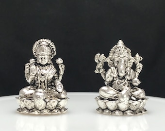 Può includere: Due piccole statuette in metallo color argento raffiguranti divinità sedute a gambe incrociate su basi a forma di fiore di loto. La figura a sinistra presenta una corona e braccia multiple, mentre quella a destra ha una testa di elefante.