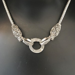 Könnte beinhalten: Eine silberne Halskette mit einem runden Anhänger und zwei blattförmigen Akzenten. Der Anhänger und die Akzente sind mit kleinen, funkelnden Steinen verziert.