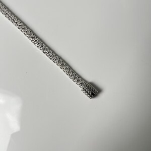 May include: A silver-colored, woven chain bracelet. The bracelet has a rectangular clasp at the end. The chain is made of small, interconnected links, creating a textured appearance. The bracelet is displayed on a white surface.