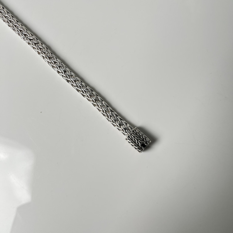 May include: A silver-colored, woven chain bracelet. The bracelet has a rectangular clasp at the end. The chain is made of small, interconnected links, creating a textured appearance. The bracelet is displayed on a white surface.