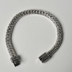 May include: A silver bracelet with a woven chain design. The bracelet is open, showing the clasp mechanism. The metal has a bright, reflective finish. The bracelet is on a white background.