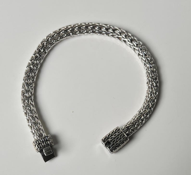 May include: A silver bracelet with a woven chain design. The bracelet is open, showing the clasp mechanism. The metal has a bright, reflective finish. The bracelet is on a white background.
