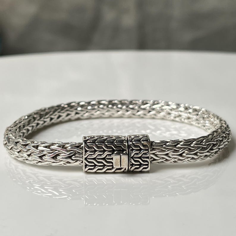 May include: A silver bracelet with a woven chain design. The bracelet features a rectangular clasp with a textured pattern. The jewelry is displayed on a white surface.