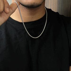 Puede incluir: Un collar de cadena de serpiente en tono plateado con un diseño de eslabones lisos, redondeados y flexibles. La cadena descansa sobre una camiseta negra lisa, mostrando una textura metálica uniforme y continua que forma un arco simple.