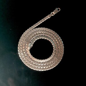 May include: A silver chain necklace with a flat, woven design. The necklace is coiled in a circular pattern against a black background, highlighting its texture and shine. The clasp is visible at the top.