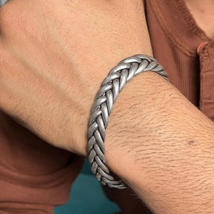 Puede incluir: Una pulsera de tono plateado con un diseño trenzado complejo de múltiples hilos y acabado metálico. La pulsera tiene un perfil ancho y plano con una superficie texturizada que imita la apariencia de eslabones metálicos tejidos en un patrón.