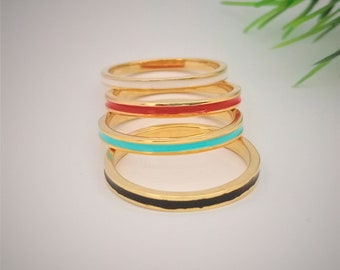 May include: Four gold rings with a thin coloured enamel band. The rings are stacked on top of each other. The top ring has a white band, the second ring has a red band, the third ring has a turquoise band, and the bottom ring has a black band.