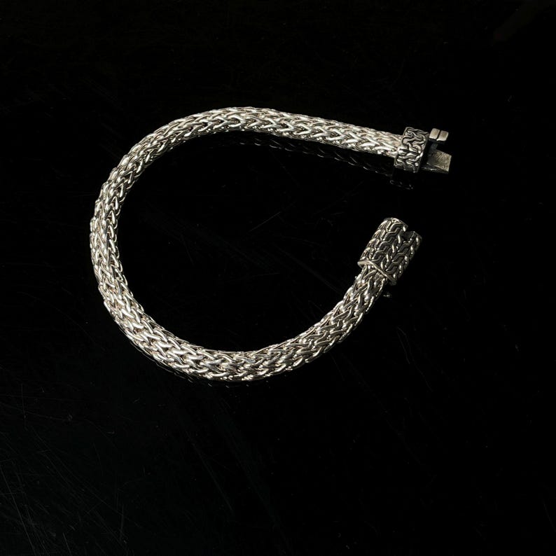 May include: A silver-colored bracelet with a woven chain design. The bracelet is unclasped, showing the clasp mechanism. The background is black, highlighting the metallic sheen of the jewelry.