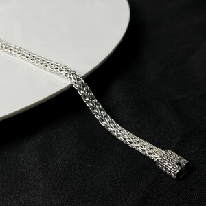 May include: A silver bracelet with a woven chain design. The bracelet is made of a shiny, metallic material and has a secure clasp. The bracelet is displayed on a white surface against a black background.