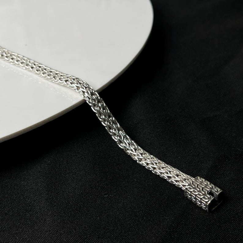 May include: A silver bracelet with a woven chain design. The bracelet is made of a shiny, metallic material and has a secure clasp. The bracelet is displayed on a white surface against a black background.