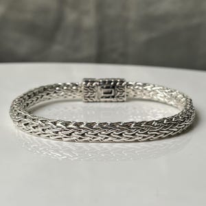 May include: A silver bracelet with a woven chain design. The bracelet features a rectangular clasp with intricate detailing. The jewelry is displayed on a white surface, with a blurred gray background.