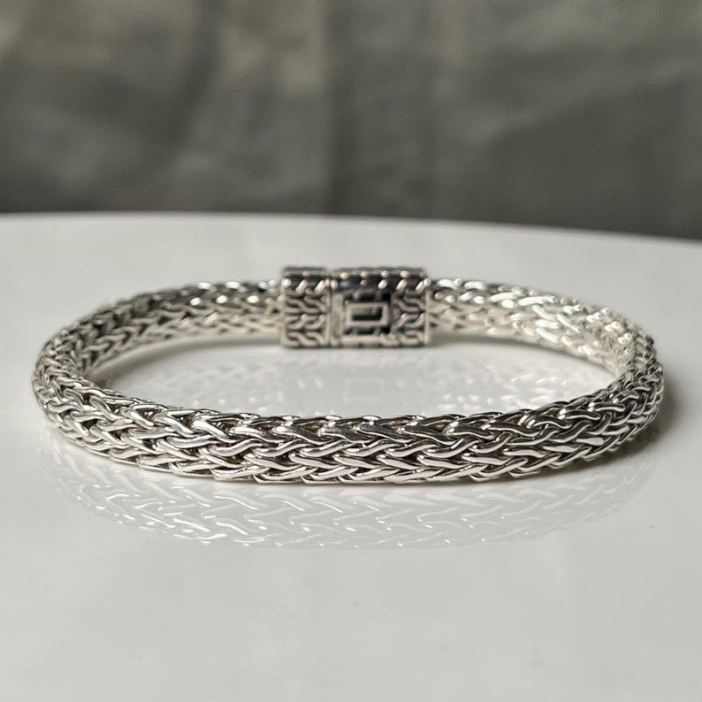 May include: A silver bracelet with a woven chain design. The bracelet features a rectangular clasp with intricate detailing. The jewelry is displayed on a white surface, with a blurred gray background.