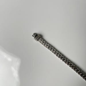 May include: A silver-toned bracelet with a woven chain design. The bracelet features a rectangular clasp with an ornate pattern. The chain is made of small, interconnected links, creating a textured surface. The bracelet is displayed against a white background.