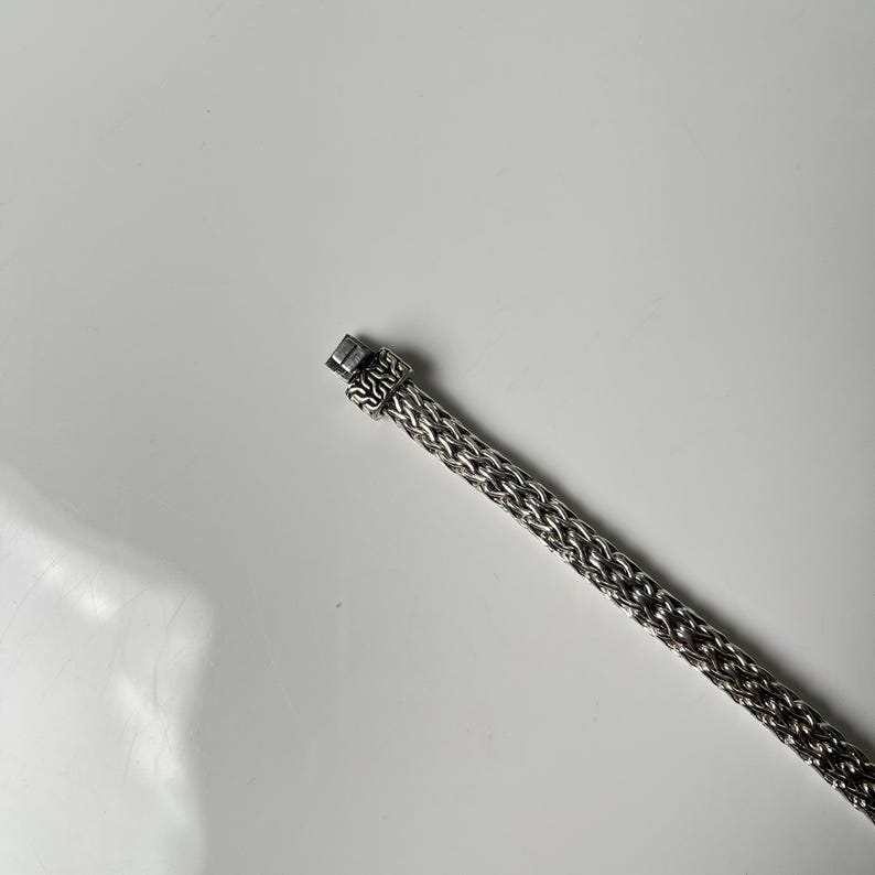 May include: A silver-toned bracelet with a woven chain design. The bracelet features a rectangular clasp with an ornate pattern. The chain is made of small, interconnected links, creating a textured surface. The bracelet is displayed against a white background.