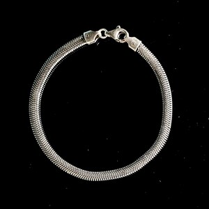 Puede incluir: Una pulsera plateada con diseño de espiga y cierre de mosquetón. La pulsera está hecha de pequeñas piezas de metal entrelazadas, creando una superficie flexible y texturizada. La pulsera se muestra sobre un fondo negro.