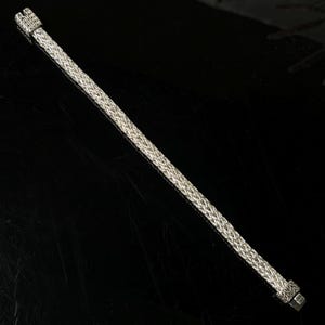 May include: A silver-colored bracelet with a woven design. The bracelet has a rectangular clasp and a textured pattern throughout its length. The bracelet is displayed on a black surface.