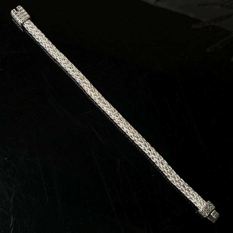 May include: A silver-colored bracelet with a woven design. The bracelet has a rectangular clasp and a textured pattern throughout its length. The bracelet is displayed on a black surface.