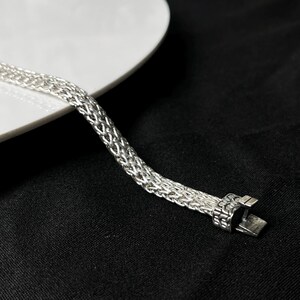May include: A silver-colored bracelet with a woven chain design. The bracelet is displayed on a black surface with a white plate in the background. The clasp is rectangular and has a secure closure.