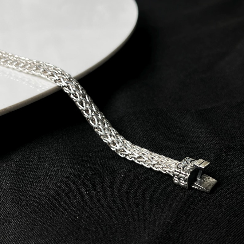 May include: A silver-colored bracelet with a woven chain design. The bracelet is displayed on a black surface with a white plate in the background. The clasp is rectangular and has a secure closure.
