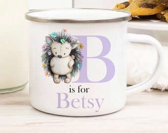 May include: White enamel mug with a silver rim, featuring a watercolor illustration of a hedgehog surrounded by flowers. A large lavender letter "B" and the text "is for Betsy" are also on the mug. A charming and personalized gift.