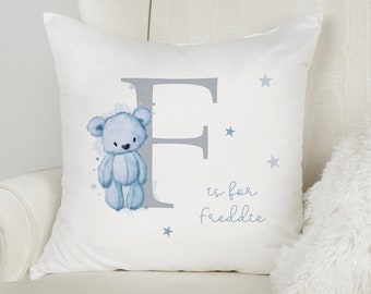 May include: White square pillow with a blue teddy bear illustration, a large gray letter "F", and the text "is for freddie" in blue. Small blue stars are scattered around the design. The pillow is on a white chair.