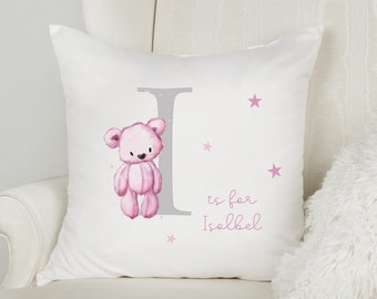 May include: White decorative pillow with a pink teddy bear illustration, a gray letter "I", and the text "is for Isabel" in pink. Pink stars are scattered around the design. The pillow is on a white chair.