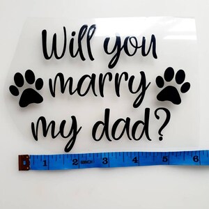 May include: Clear, rectangular heat transfer vinyl with the text "Will you marry my dad?" in black script, flanked by two black paw prints. A blue measuring tape shows the width is approximately 6 inches.