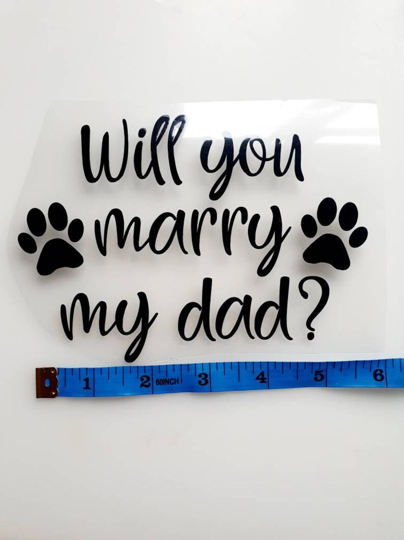 May include: Clear, rectangular heat transfer vinyl with the text "Will you marry my dad?" in black script, flanked by two black paw prints. A blue measuring tape shows the width is approximately 6 inches.