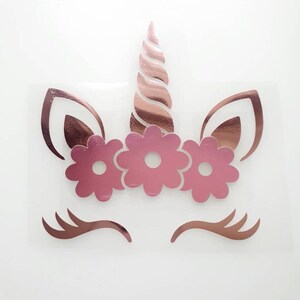 May include: A rose gold unicorn face design with a horn, ears, eyelashes, and a floral crown. The unicorn design is on a clear background. The design is a sticker or iron-on transfer.