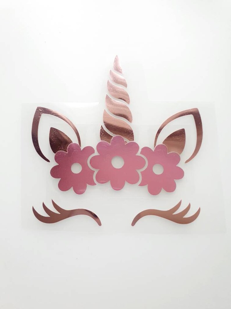 May include: A rose gold unicorn face design with a horn, ears, eyelashes, and a floral crown. The unicorn design is on a clear background. The design is a sticker or iron-on transfer.