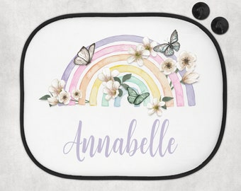 May include: White sunshade featuring a watercolor rainbow with butterflies and flowers. The name "Annabelle" is written in a soft purple script. Includes two black suction cups for attachment.