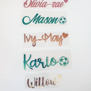 May include: Five personalized name decals in various colors. The names are in cursive script, with heart or soccer ball accents. Names include Olivia-rae, Mason, Ivy-May, Kario, and Willow. The decals are on a clear backing.