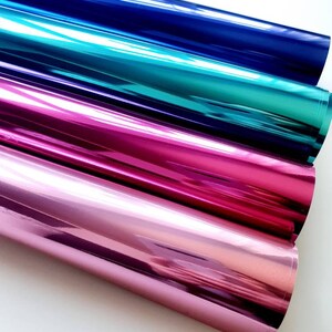 May include: A close-up of four rolls of shiny, metallic foil in shades of blue, teal, pink, and rose. The foil reflects light, creating a glossy appearance. The rolls are stacked next to each other, with the edges visible.
