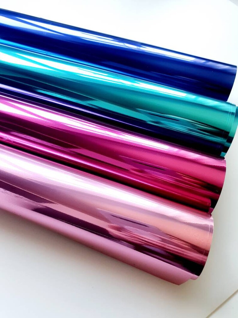 May include: A close-up of four rolls of shiny, metallic foil in shades of blue, teal, pink, and rose. The foil reflects light, creating a glossy appearance. The rolls are stacked next to each other, with the edges visible.