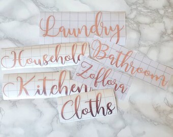 May include: Rose gold vinyl labels with cursive text for home organization. Labels include "Laundry", "Household", "Kitchen", "Bathroom", "Zoflora", and "Cloths". The labels are on a grid backing, set against a marble background.