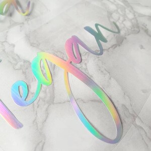 May include: Close-up of iridescent vinyl lettering spelling out the name "Megan" in a cursive font. The letters shift colors, displaying hues of pink, green, and blue. The vinyl is on a clear backing, with a marble-patterned surface in the background.