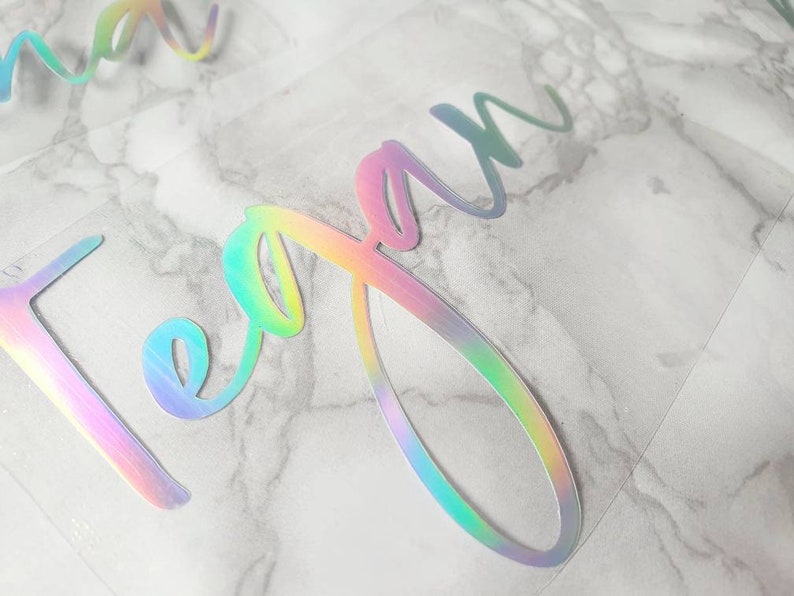 May include: Close-up of iridescent vinyl lettering spelling out the name "Megan" in a cursive font. The letters shift colors, displaying hues of pink, green, and blue. The vinyl is on a clear backing, with a marble-patterned surface in the background.