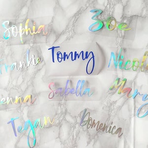 May include: A selection of personalised name decals in iridescent and blue hues. Names such as "Sophia", "Tommy", and "Nicole" are written in a cursive script. The decals are on a transparent backing, presented against a marble-effect backdrop.