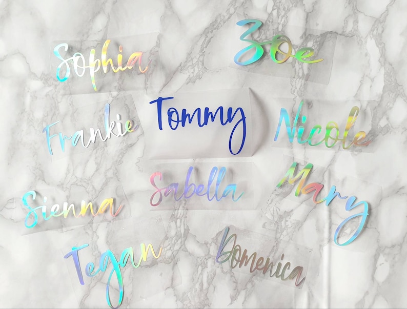 May include: A collection of custom name decals in various colors, including iridescent and blue. Names like "Sophia", "Tommy", and "Nicole" are displayed in a cursive font. The decals are on a clear backing, set against a marble-patterned background.