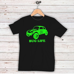 Może przedstawiać: Czarny t-shirt z neonowo-zieloną grafiką samochodu i napisem "BUG LIFE" poniżej. Samochód ma numer "01". Koszulka wisi na drewnianym wieszaku na tle jasnego drewnianego tła.