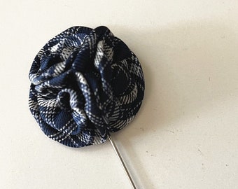 May include: A navy blue and white plaid fabric flower lapel pin with a long silver pin. The fabric is arranged in a circular shape, creating a textured, layered effect. The pin is a stylish accessory.
