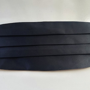 May include: A navy blue cummerbund with a pleated design. The formal accessory is made of a smooth, satin-like fabric. It is designed to be worn around the waist with formal attire.
