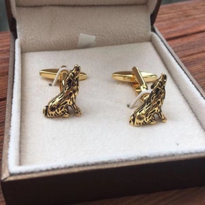 May include: A pair of gold cufflinks featuring a wolf design. The wolf is howling and sitting on its haunches. The cufflinks are in a brown box.