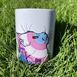 Puede incluir: Taza de cerámica blanca con un diseño de zorro estilizado. La cara del zorro está decorada con patrones azules, rosas y morados. La taza es cilíndrica y está sobre hierba verde, bajo la luz del sol.