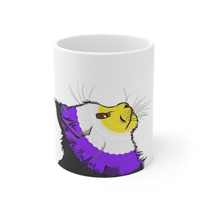 Op de afbeelding: Witte keramische mok met een gestileerd kattenontwerp in de kleuren van non-binaire trots. De kat heeft een gele kop, met paars en zwart haar. Het ontwerp is gedrukt op de zijkant van de mok.
