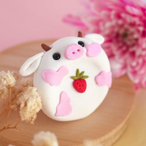 May include: A round white clay figurine shaped like a cow with pink spots, a pink snout, small brown horns, and black eyes. A small red strawberry with green leaves is attached to the center of the cow's body. The figurine has two white ears.