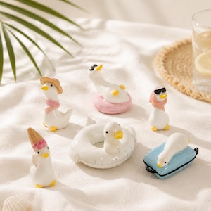 May include: A collection of six small ceramic duck figurines. One wears a straw hat and pink scarf, one floats in a pink ring, one wears sunglasses and a pink scarf, one wears an ice cream cone hat, one sits in a white ring, and one lies on a blue suitcase.