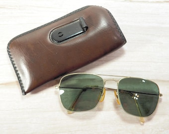 May include: A pair of gold-framed sunglasses with green lenses rests beside a brown leather case. The case has a black clasp and a stitched border. The sunglasses have a classic aviator design.