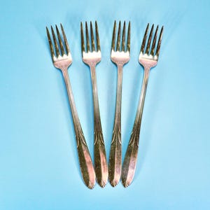 May include: A set of four silver-toned metal forks arranged vertically. Each fork features a four-tine head and a slender handle decorated with a series of vertical, embossed lines that taper toward the base of the handle, creating a uniform design.